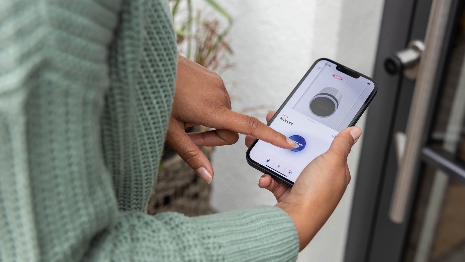Main ouvrant une porte d'entrée depuis l'application ABUS One affichée à l'écran d'un smartphone