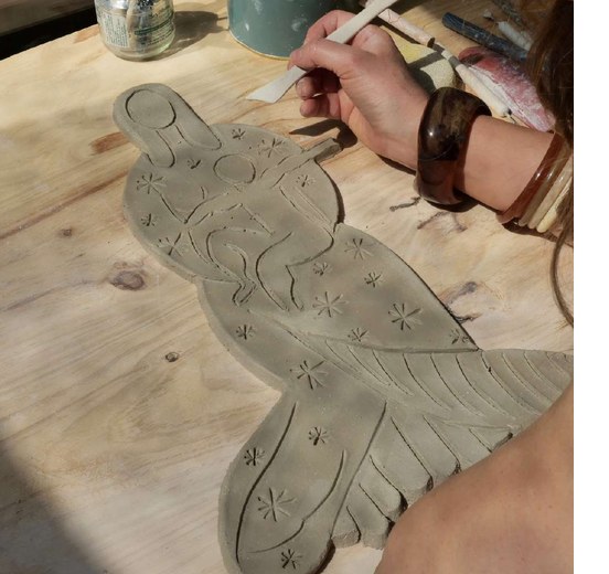 Main tenant un outil pour graver les motifs d'un oiseau dans une pièce de terre crue, sur un établi