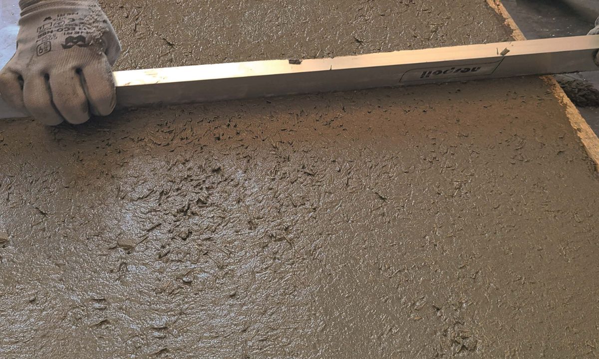 Tirage à la règle d'une chape fraîche sur un plancher, mains gantées tenant une règle alu
