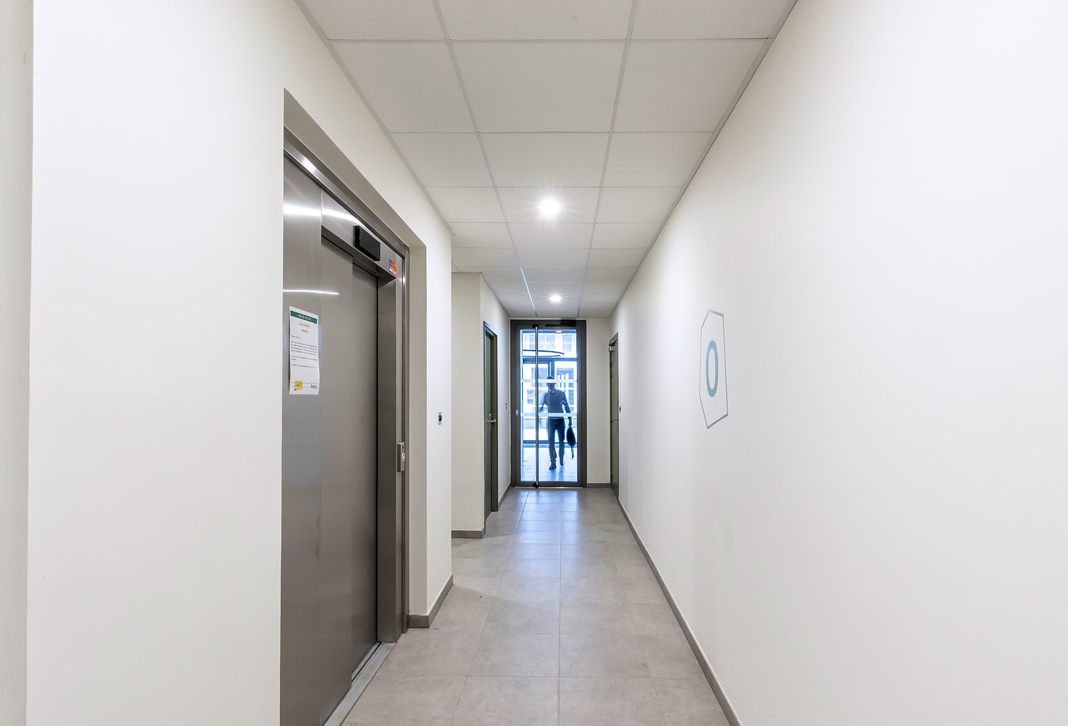 Couloir en enfilade d'un immeuble résidentiel, plafond modulaire acoustique et porte d'ascenseur en inox.