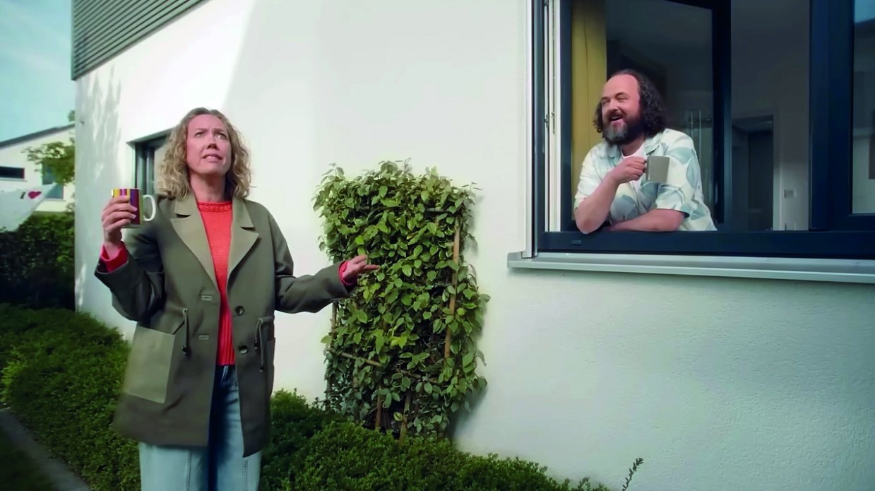 La voisine Louise dans le jardin et Hugo riant à sa fenêtre, scène du spot VEKA Hugo et sa voisine