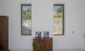 Deux fenêtres aluminium étroites Cuzco Jaya dans un mur blanc, de part et d’autre d’une commode ancienne