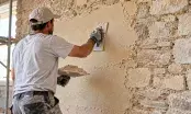 Applicateur talochant un enduit à la chaux sur un mur en pierre calcaire, moellons apparents à droite