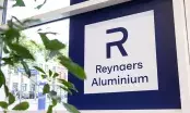 Enseigne Reynaers Aluminium fixée derrière une façade vitrée, feuillage vert au premier plan
