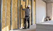 Artisan plaçant un panneau de laine minérale entre les montants métalliques d'une contre-cloison, plaques de plâtre empilées
