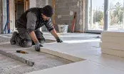 Un artisan agenouillé pose des panneaux isolants rigides sur la dalle béton d’une maison en rénovation