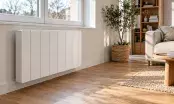 Radiateur électrique blanc fixé sous une fenêtre dans un séjour clair à parquet chêne, lumière naturelle d'hiver