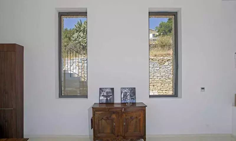 Deux fenêtres aluminium étroites Cuzco Jaya dans un mur blanc, de part et d’autre d’une commode ancienne