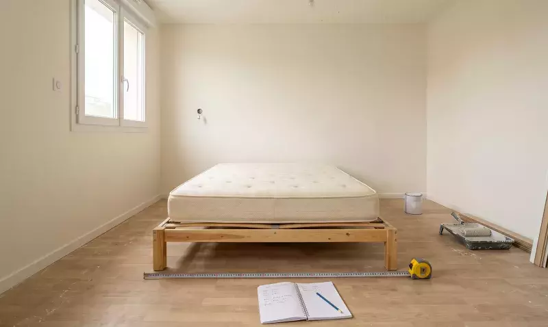 Chambre d'un logement en rénovation avec un lit deux places, mètre à ruban déroulé au sol le long du couchage