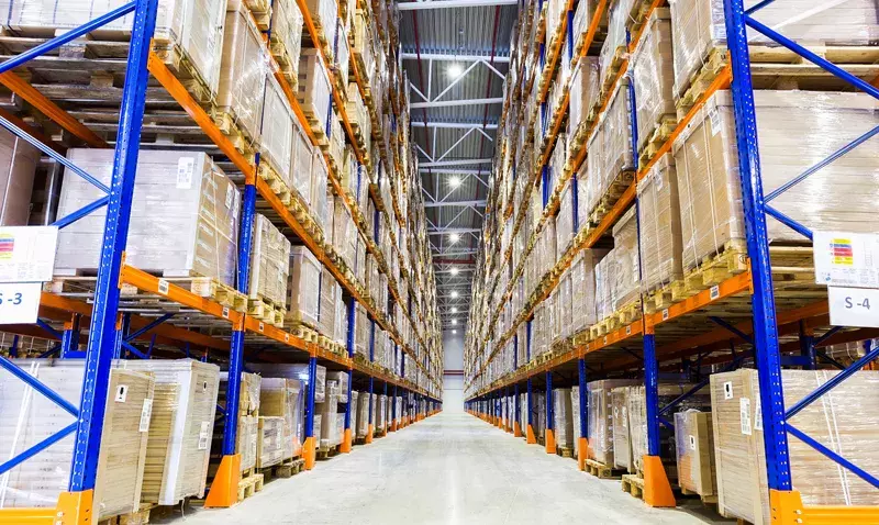 Allée d'entrepôt logistique bordée de rayonnages hauts, éclairée par une ligne de luminaires LED fixés en plafond.
