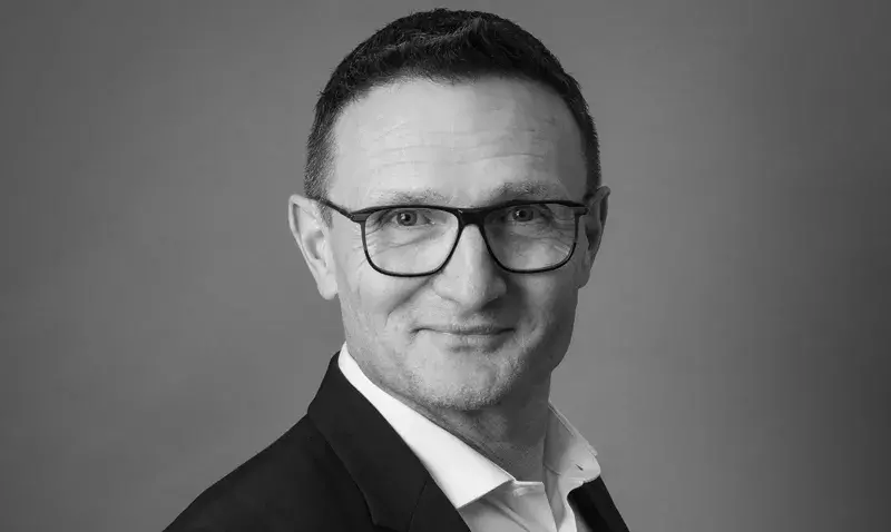 Portrait en noir et blanc d'un homme souriant, cheveux courts et lunettes, en costume, sur fond gris uni de studio.