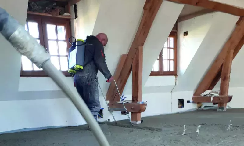 Chapiste réglant une chape fraîchement pompée dans un comble aménagé, le tuyau de pompe traversant la pièce