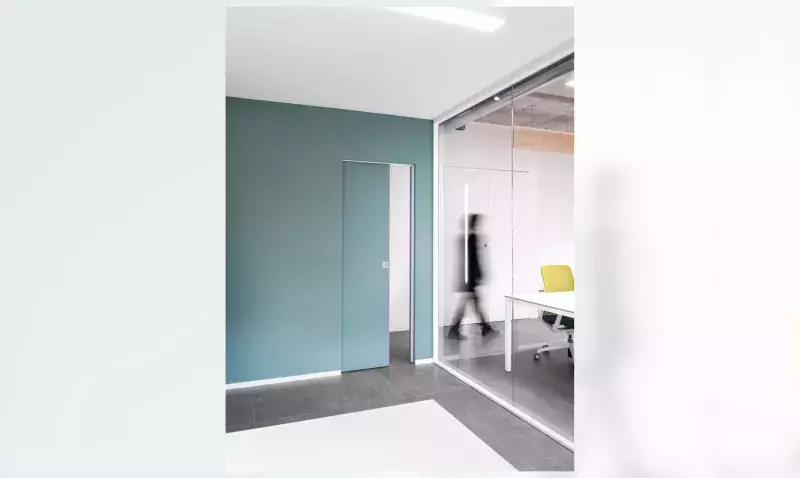Porte coulissante affleurante dans un mur bleu-vert d'open space, cloison vitrée et sol gris