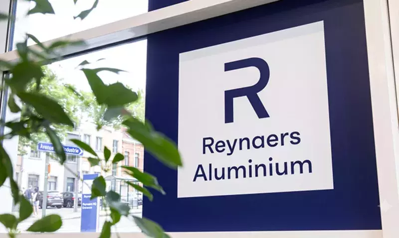 Enseigne Reynaers Aluminium fixée derrière une façade vitrée, feuillage vert au premier plan