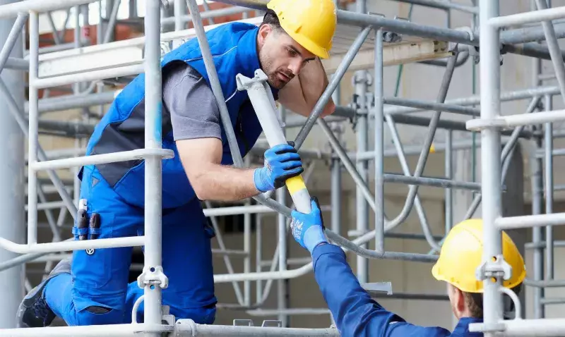 Deux ouvriers portant des gants de protection bleus assemblent les tubes métalliques d'un échafaudage de chantier