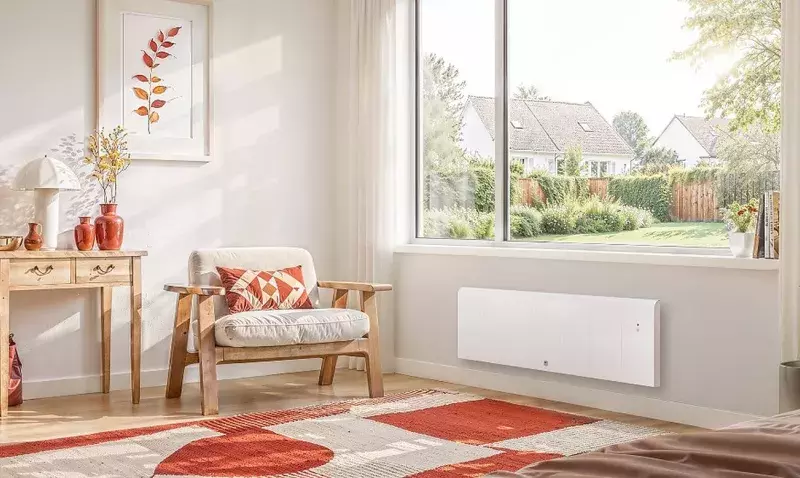 Radiateur à chaleur douce au format plinthe posé sous une fenêtre de chambre, près d'un fauteuil en bois.