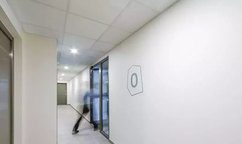 Couloir d'immeuble de logements équipé d'un plafond modulaire à dalles blanches, avec ascenseur et une personne qui passe.