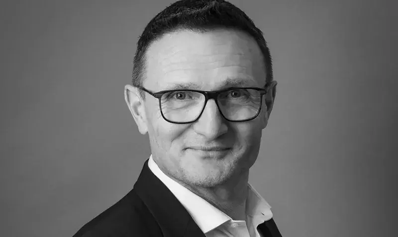Portrait en noir et blanc d'un homme souriant, cheveux courts et lunettes, en costume, sur fond gris uni de studio.