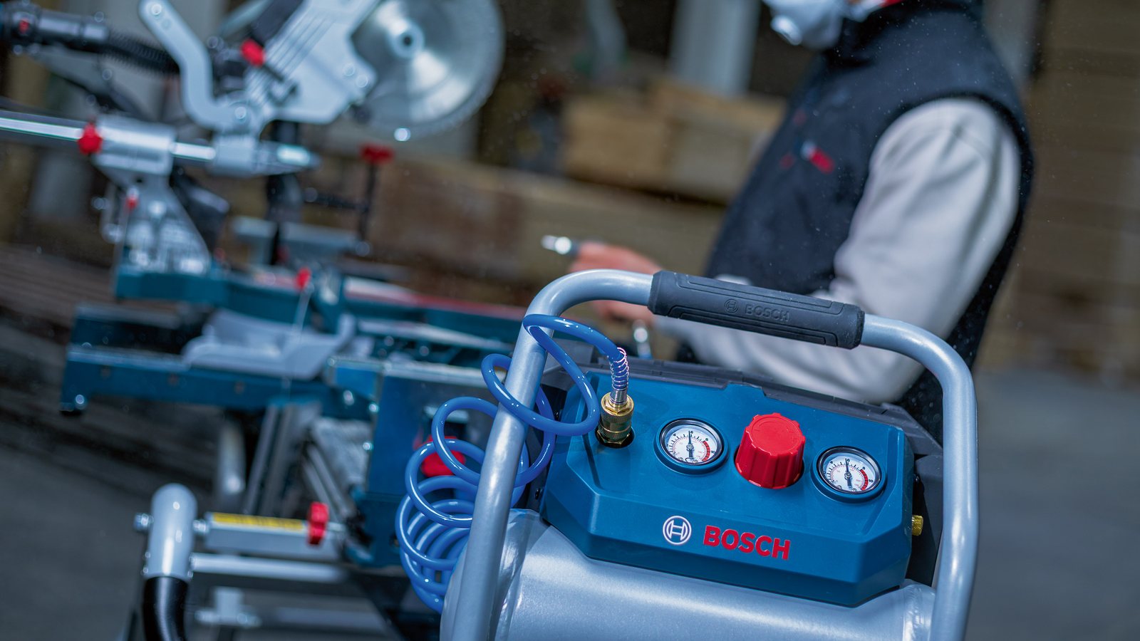 Compresseur Bosch GKO18V-51 au premier plan dans un atelier bois, scie à onglet radiale à l'arrière-plan